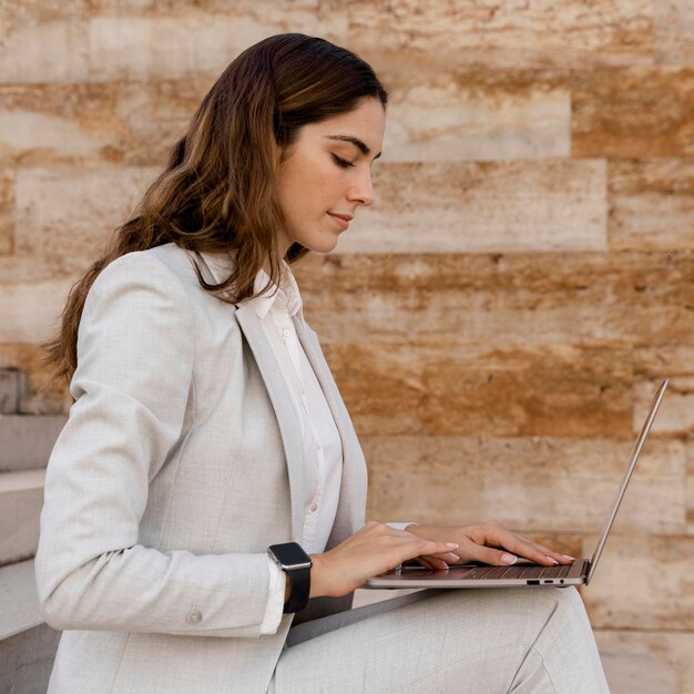 Бесплатное фото Вид сбоку элегантной бизнес-леди с умными часами, работающих на ноутбуке на открытом воздухе