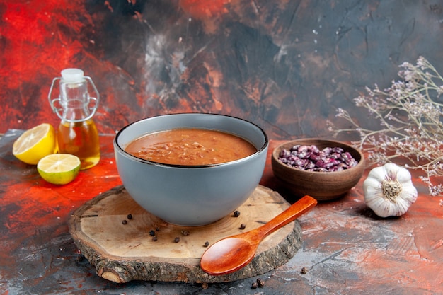 Бесплатное фото Вид сбоку томатный суп с ложкой на деревянном подносе, бутылка масла из бобов и чеснок, лимон, помидор на столе смешанных цветов