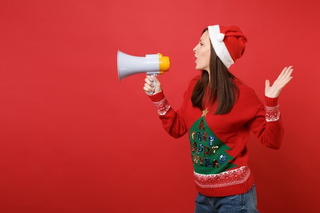 빨간 배경에 격리된 확성기를 들고 있는 크리스마스 모자, 니트 스웨터를 입은 어린 산타 소녀의 측면. 새해 복 많이 받으세요 2019 축 하 휴일 파티 개념입니다. 복사 공간을 비웃습니다.