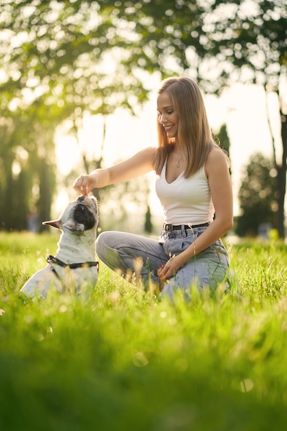 都市公園でフレンチブルドッグを訓練している若い笑顔の女性の側面図。純血種のペットの匂いが女性の犬の飼い主の手からおやつ、背景に美しい夏の夕日。動物調教の概念。