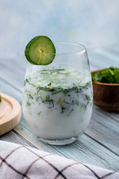 Side view of okroshka in a glass with a cucumber and a white checkered towel