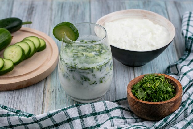 Side view of okroshka in a glass with cucumbers on a stand yogurt and herbs on a gray surface