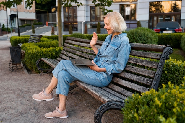 Free photo side view of older woman outdoors on bench waving at laptop