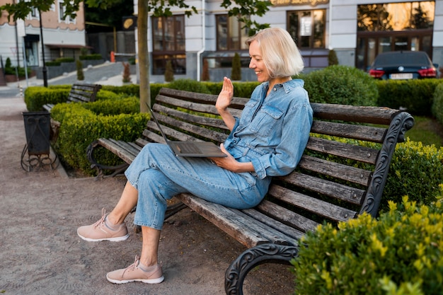 Side view of older woman outdoors on bench waving at laptop