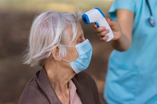 彼女の温度をチェックしている医療マスクを持つ年上の女性の側面図