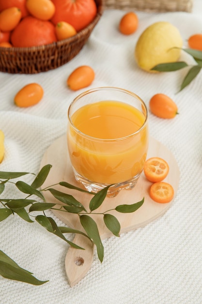 Side view of orange juice in glass with kumquat slices on cutting board and tangerines kumquats lemon with leaves on white cloth background