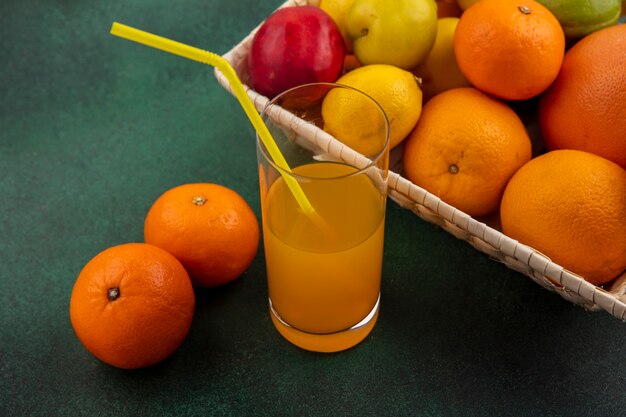 Вид сбоку апельсиновый сок в стакане с апельсинами, лимонами и алычой в корзине на зеленом фоне