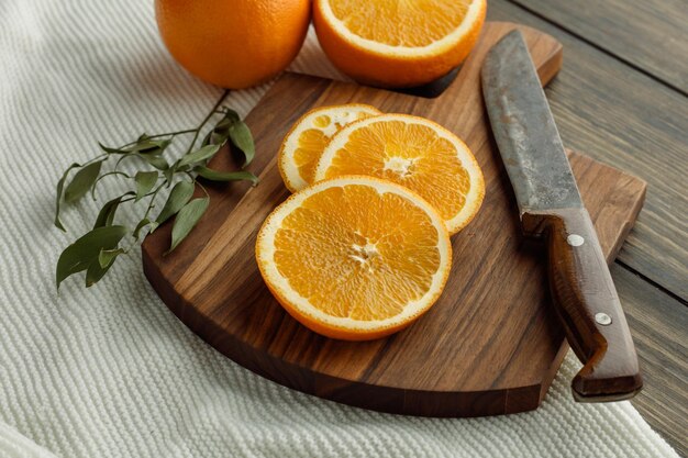 Side view of orange slices with knife on cutting board with whole and half cut orange and leaves on cloth on wooden background
