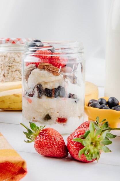 Side view of overnight oats with fresh strawberries blueberries and nuts in a glass jar on rustic table