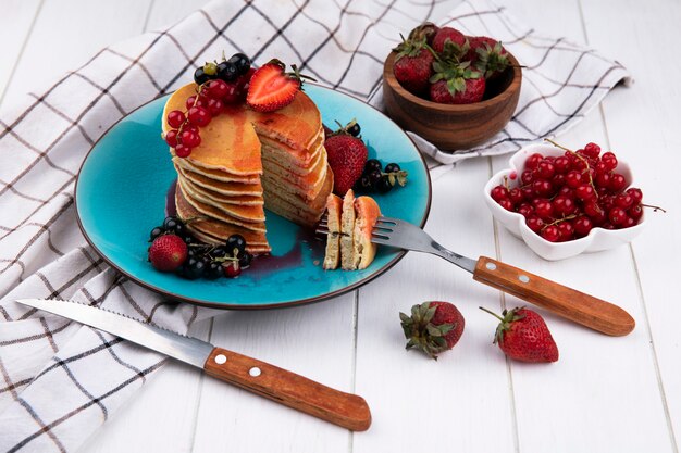 Side view pancakes with black and red currants strawberries with a fork and a knife on a plate on a white checkered towel