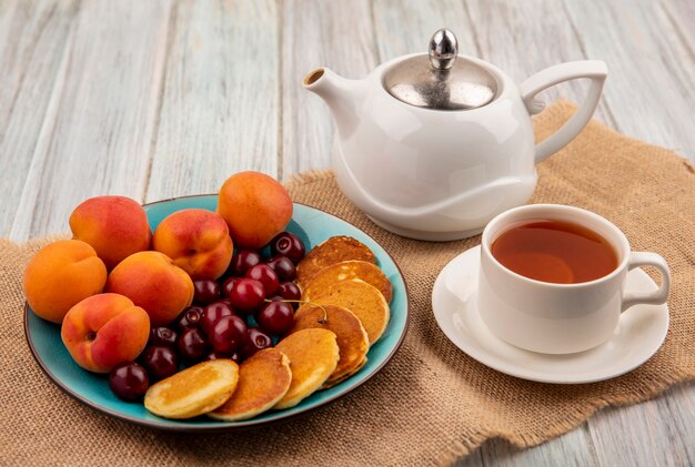 Side view of pancakes with cherries and apricots in plate and cup of tea with teapot on sackcloth and wooden background