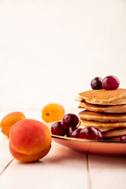 Side view of pancakes with cherries in plate and apricot on wooden surface and white background