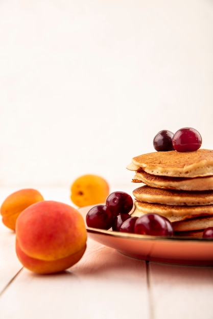 Side view of pancakes with cherries in plate and apricot on wooden surface and white background