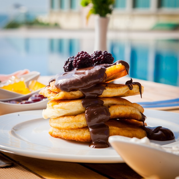 Side view pancakes with chocolate and blackberry on a wooden table against the surface of the pool