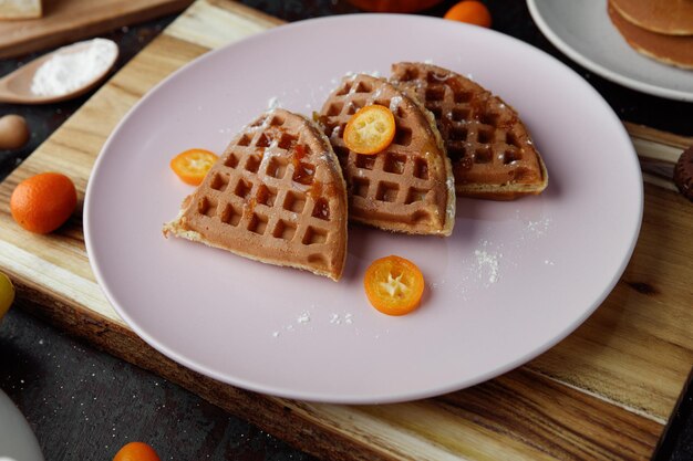 Side view of pancakes with kumquat slices in plate and kumquat on cutting board with spoonful of flour on black background