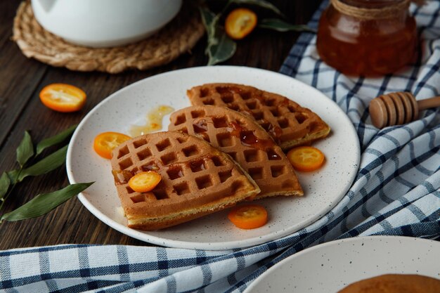 Side view of pancakes with kumquat slices in plate with jam on plaid cloth on wooden background