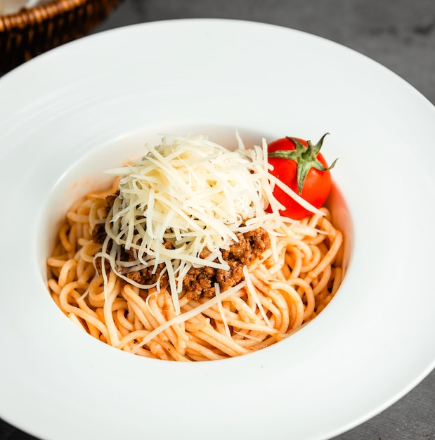Side view of pasta with minced meat grated cheese and fresh tomato in white plate on black
