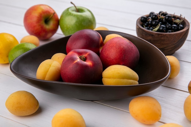 Side view of peaches with apricots in a bowl with black currant on a white surface