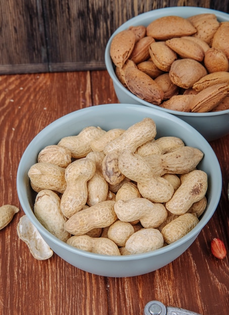 Side view of peanuts in shell in a bowl with almond on wooden background