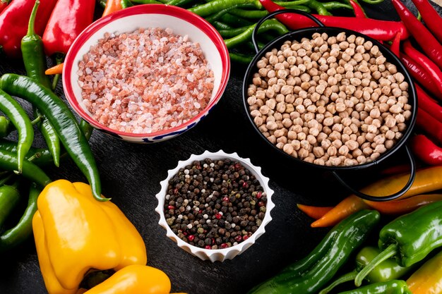 Side view pepper chili bell chickpeas in black pan and spices in white bowls on dark stone