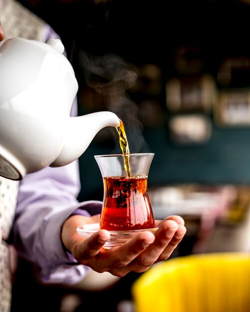 Side view of a person pouring black tea from white ceramic teapot into armudu glass