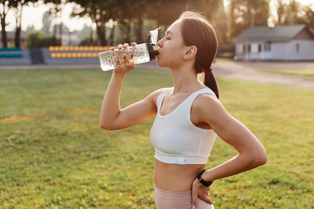 Side view photo of dark haired woman wearing white top drinking water from the bottle, keeps hand on hip, feeling thirsty during training outdoor, healthy lifestyle.