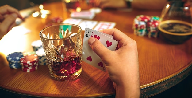 Side view photo of male and female friends sitting at wooden table. Men and women playing card game. Hands with alcohol close-up.
