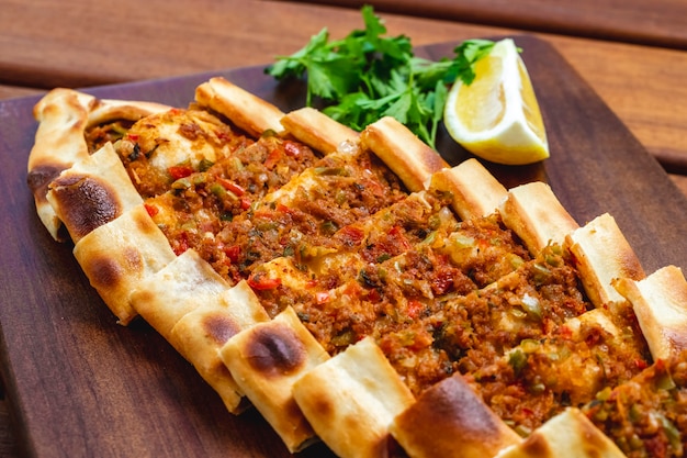 Side view pide with ground meat tomato onion hot green pepper greens and slice of lemon on a tray