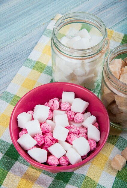 Side view of pink sugar candies in a bowl and different types of sugar in glass jars on plaid table napkin on rustic background