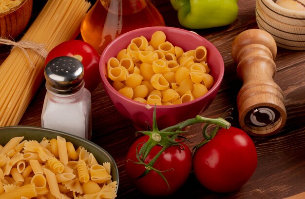 Side view of pipe-rigate pasta in bowl with other types in another bowl and salt tomato butter pepper on wooden table