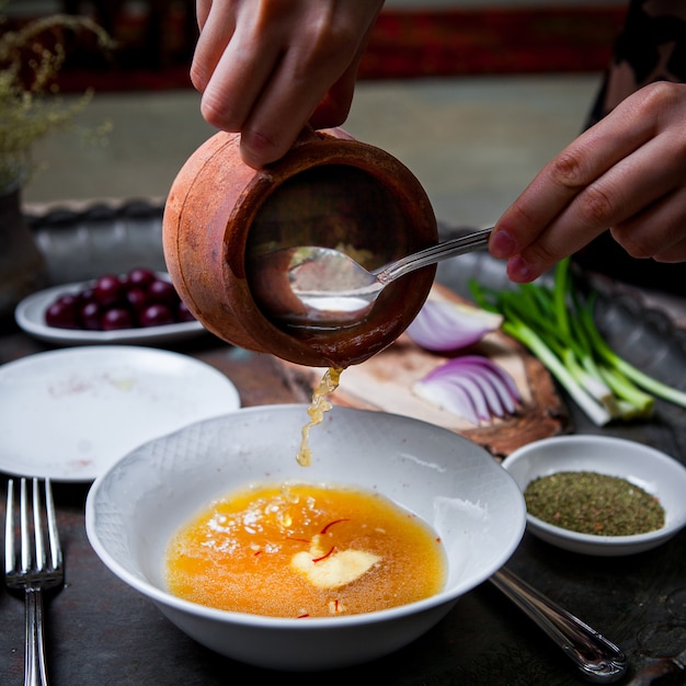 Side view piti in clay pot with deep plate and green onions and human hand and spoon in copper tray on restaurant