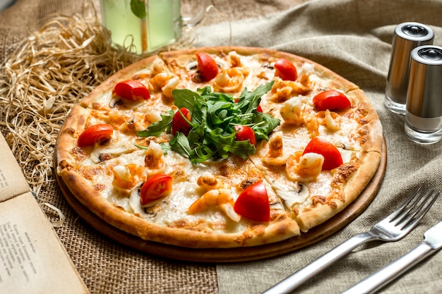 Pizza vista laterale con gamberi e funghi pomodori e rucola e con una bibita analcolica