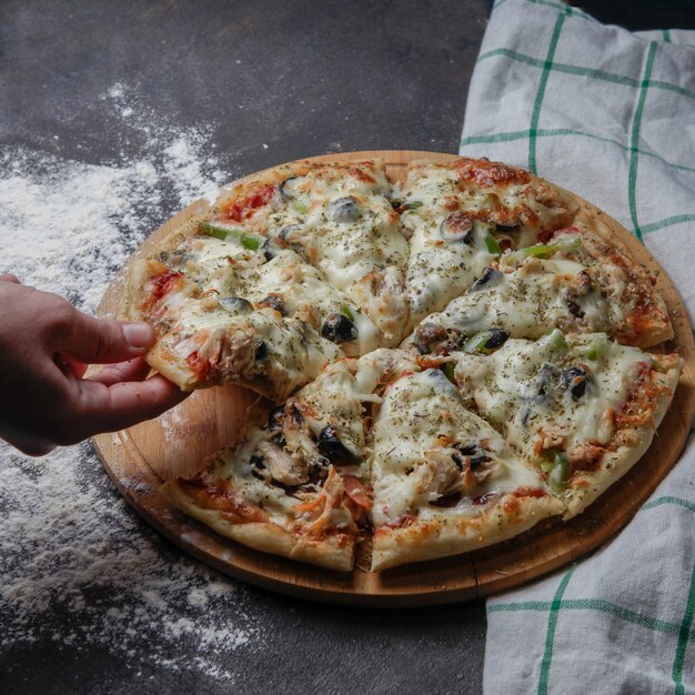 Вид сбоку пиццы на деревянной подставке со скатертью, рукой взять кусок пиццы