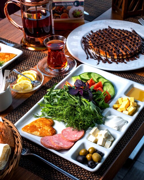 Side view of a plate with breakfast food fresh vegetable salad cheese honey fried eggs and sausages served with tea and desert