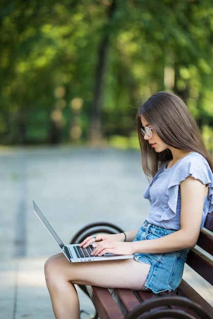 公園のベンチに座って、ラップトップコンピューターを使用して眼鏡をかけて満足しているブルネットの女性の側面図