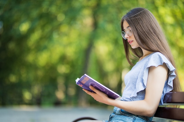 Side view of pleased brunette woman in eyeglasses sitting on bench and reading book in park