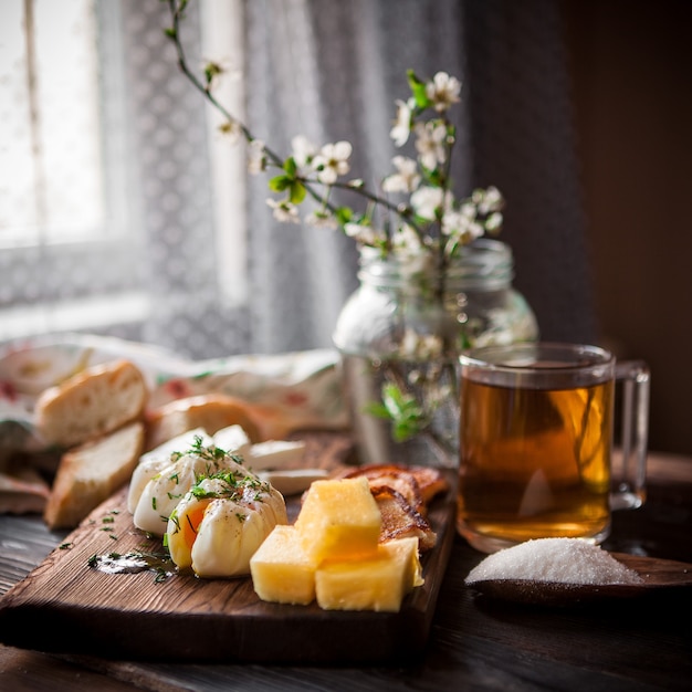 Side view poached egg with cup of tea and cheese and flowers in jar in board cookware