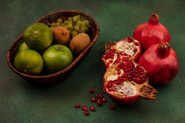 Foto gratuita metà del melograno di vista laterale con l'uva della mela e il kiwi dei mandarini in un canestro su una parete verde