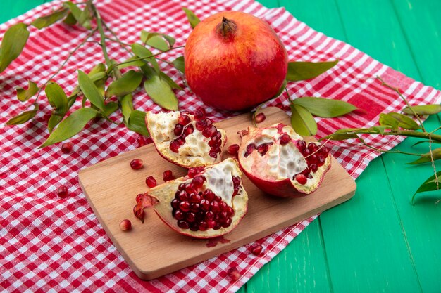 Side view of pomegranate pieces on cutting board with whole one and leaves on plaid cloth and green
