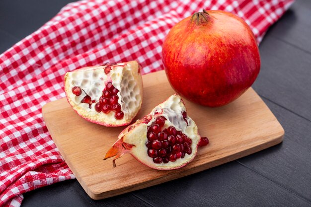Side view of pomegranate pieces and whole one on cutting board on plaid cloth and black