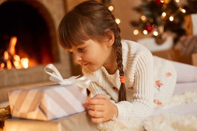 Портрет вида сбоку милый ребенок женского пола в белом свитере и шляпе Санта-Клауса, позирующий в праздничной комнате с камином и рождественской елкой, играя возле подарочных коробок на Новый год.