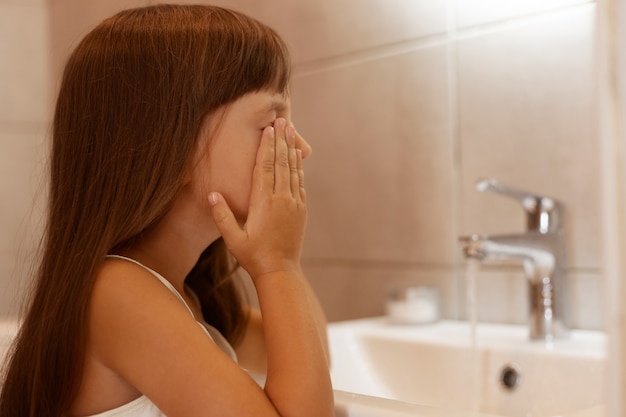 Side view portrait of dark haired female child washing her face in bathroom after waking up in the morning, standing in bathroom near faucet, and sink.