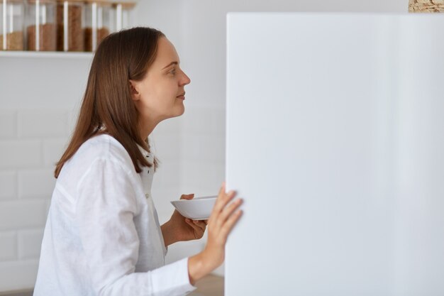 Side view portrait of dark haired woman looking for something in the fridge at home, standing with plate in hands, wearing white shirt, feels hungry, finds food.