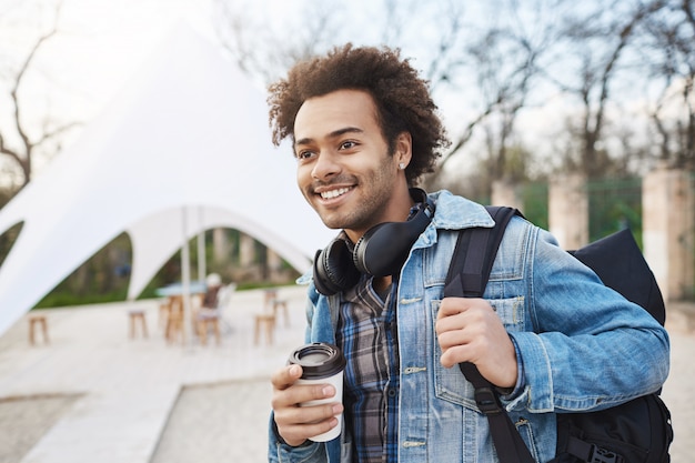 Free photo side-view portrait of fashionable happy dark-skinned guy holding backpack and coffee while walking in city, smiling and looking aside, being in good mood