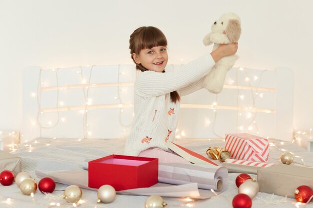 Side view portrait of little girl wearing white sweater holding soft toy dog, sitting on bed with Christmas decoration and garland, raised arms and showing her New year present.