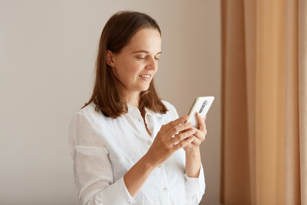 Портрет вид сбоку позитивной женщины с темными волосами в белой хлопковой рубашке, стоящей с мобильным телефоном в руках возле окна в гостиной дома, просматривая Интернет.