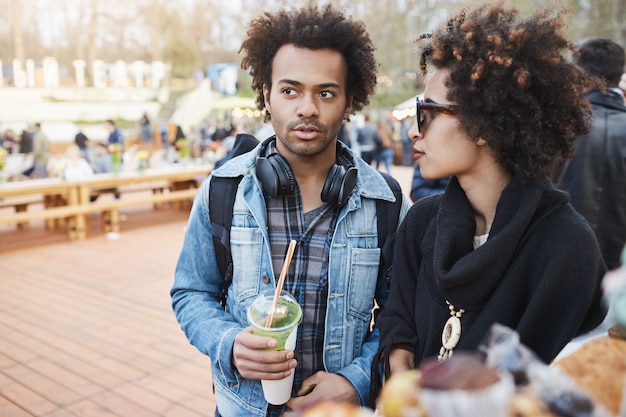ガールフレンドとフードフェスティバルの上を歩いて、コーヒーを飲みながら話しているアフロの髪型と深刻な魅力的な浅黒いボーイフレンドのサイドビューの肖像画