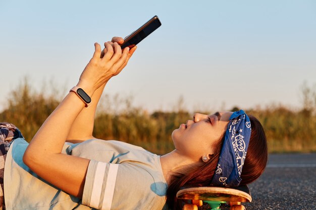 Side view portrait of slim beautiful woman wearing hair band and t shirt laying on asphalt road and keeping head on skateboard, holding phone in hands, making selfie or browsing internet.