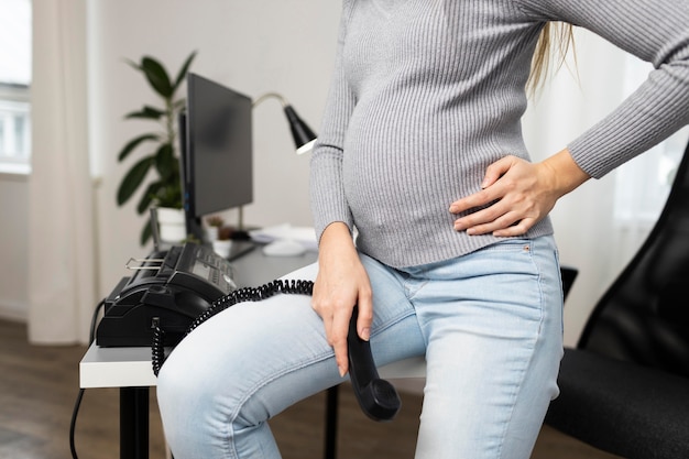 Side view of pregnant businesswoman sitting on desk and holding phone receiver
