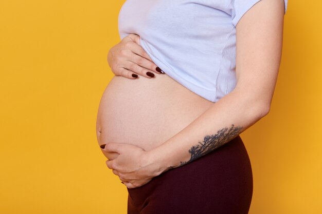 Side view of pregnant female with naked belly isolated over yellow wall. Woman with tattoo on hend dressed casually being photographed. Motherhood and pragnancy concept.